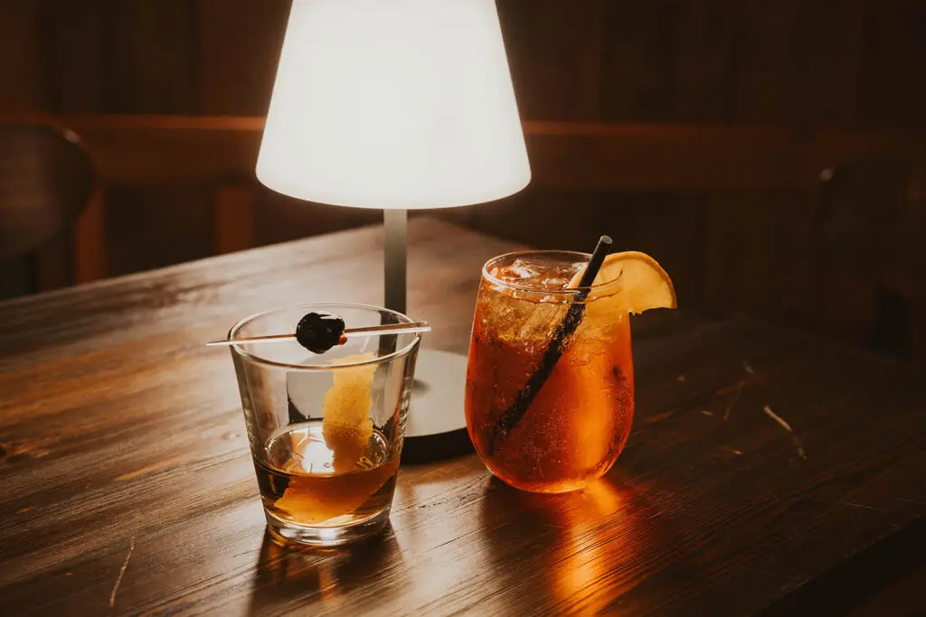 Two cocktails in glasses sitting on a wooden table with a moody lamp in the bakcdrop.