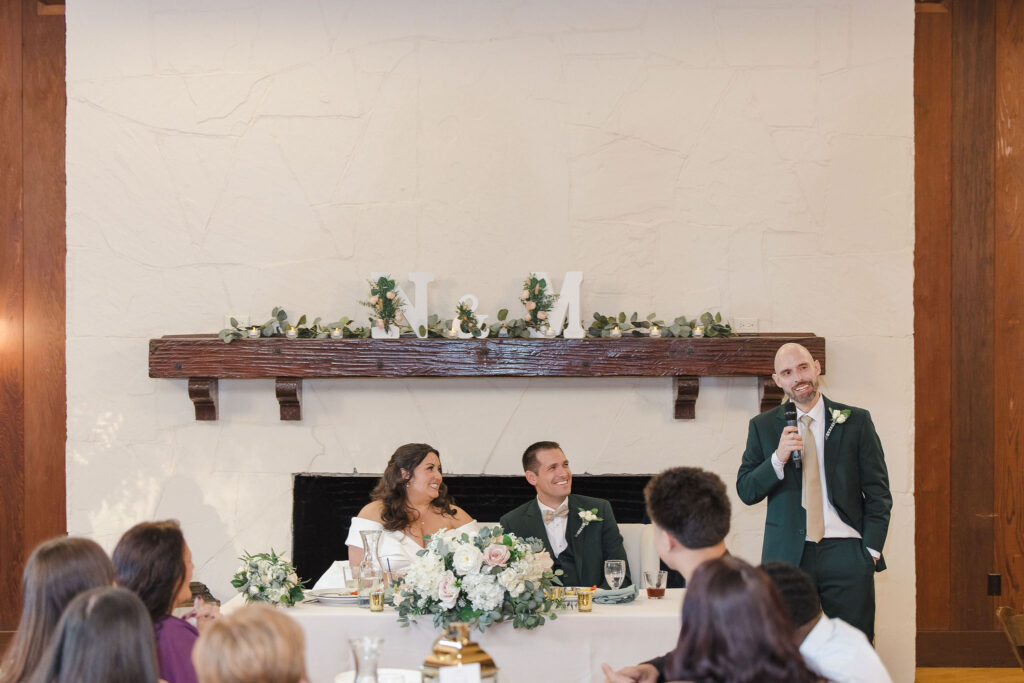 Bride and groom sitting at sweetheart table while groomsman stands next to the table delivering a speech with microphone.